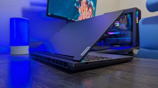 A Lenovo Legion 5i (Gen 9) gaming laptop on a desk with a blue backlight, facing away at an angle with its lid open to show its rear and right side ports, a bit of its RGB-lit keyboard, and the "Lenovo" logo on the lid.